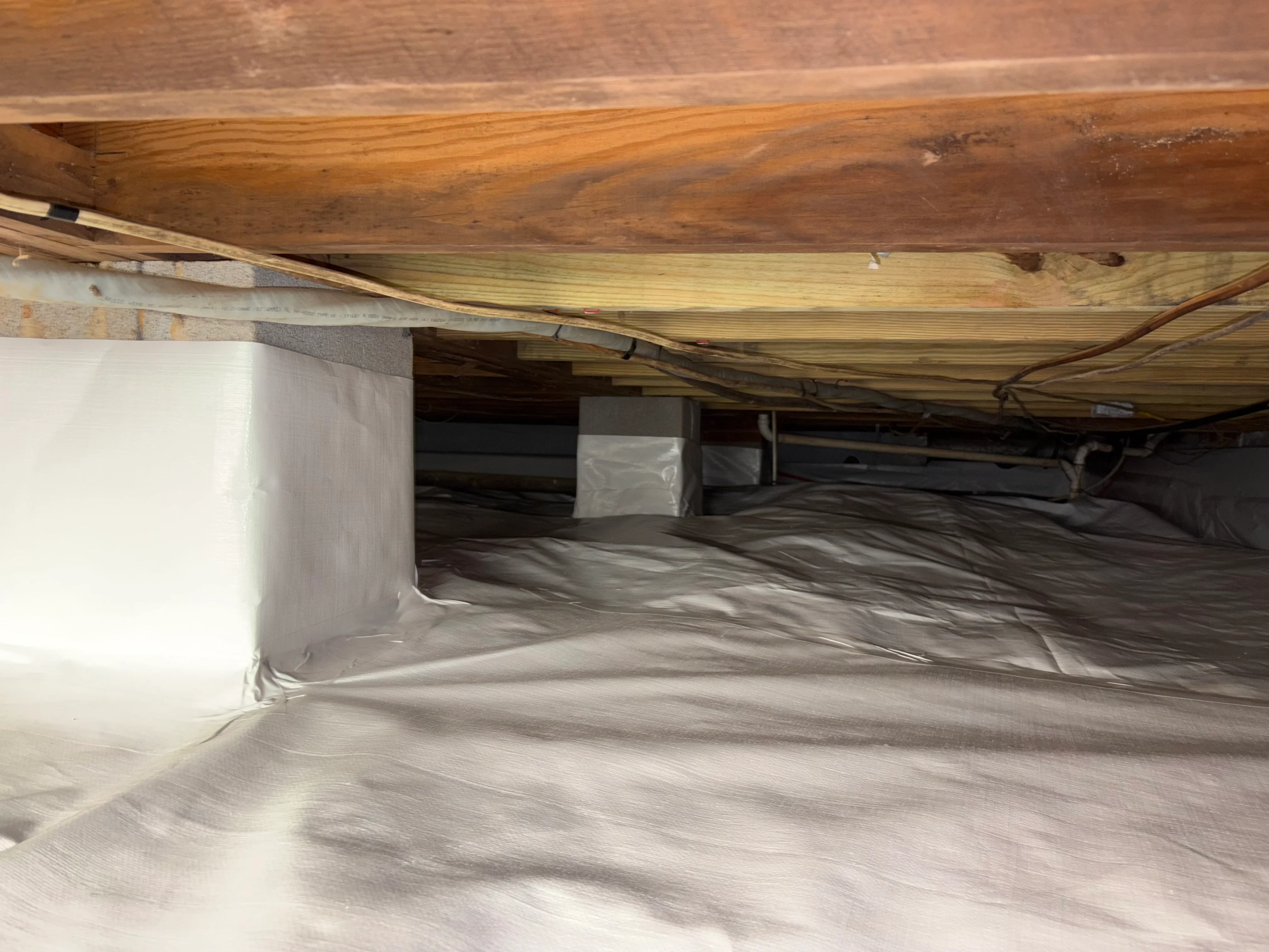 Crawl space with white vapor barrier and wooden joists visible overhead