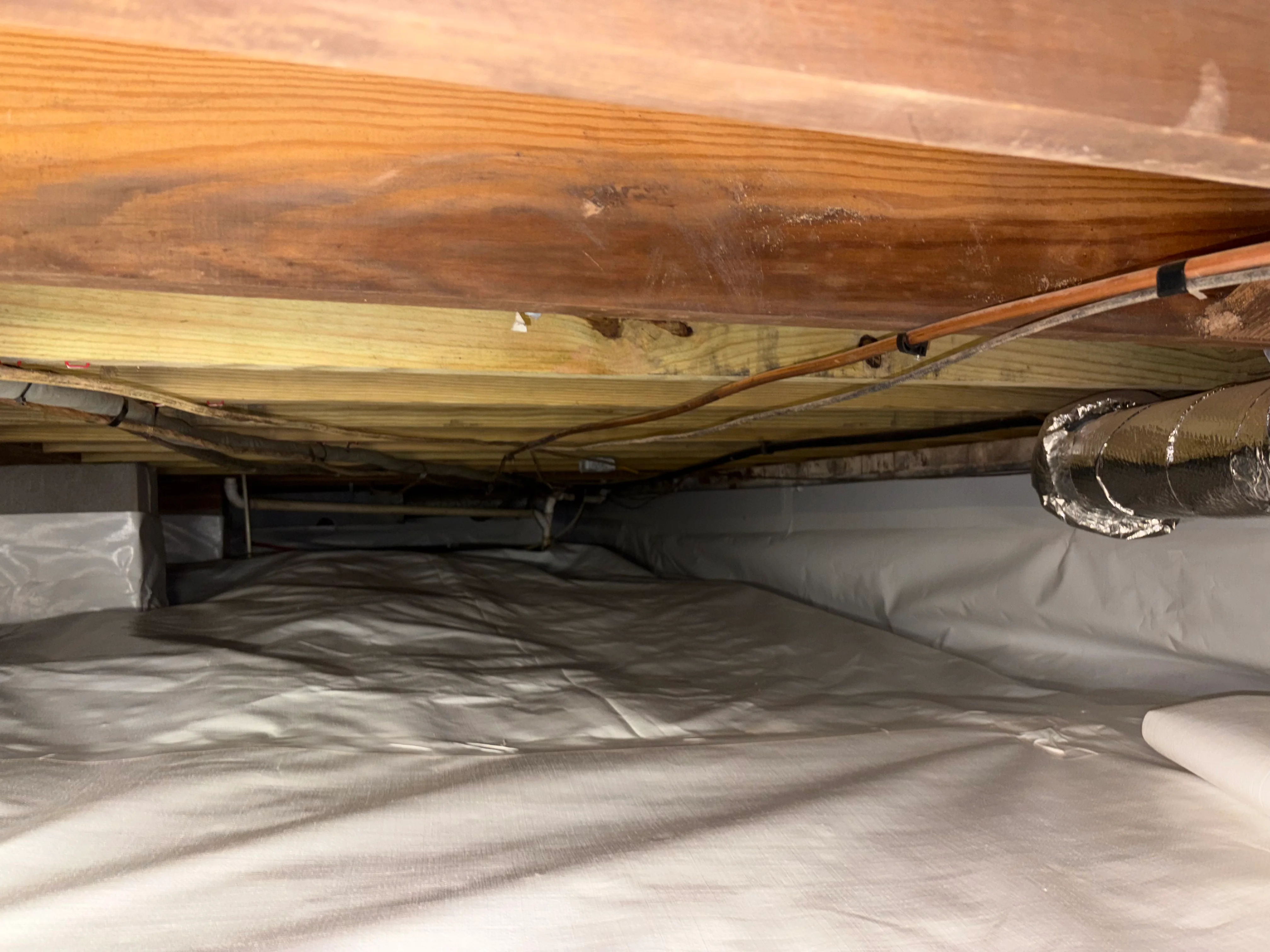 Crawl space with white vapor barrier, foam insulation, and sealed HVAC ducts under wooden joists