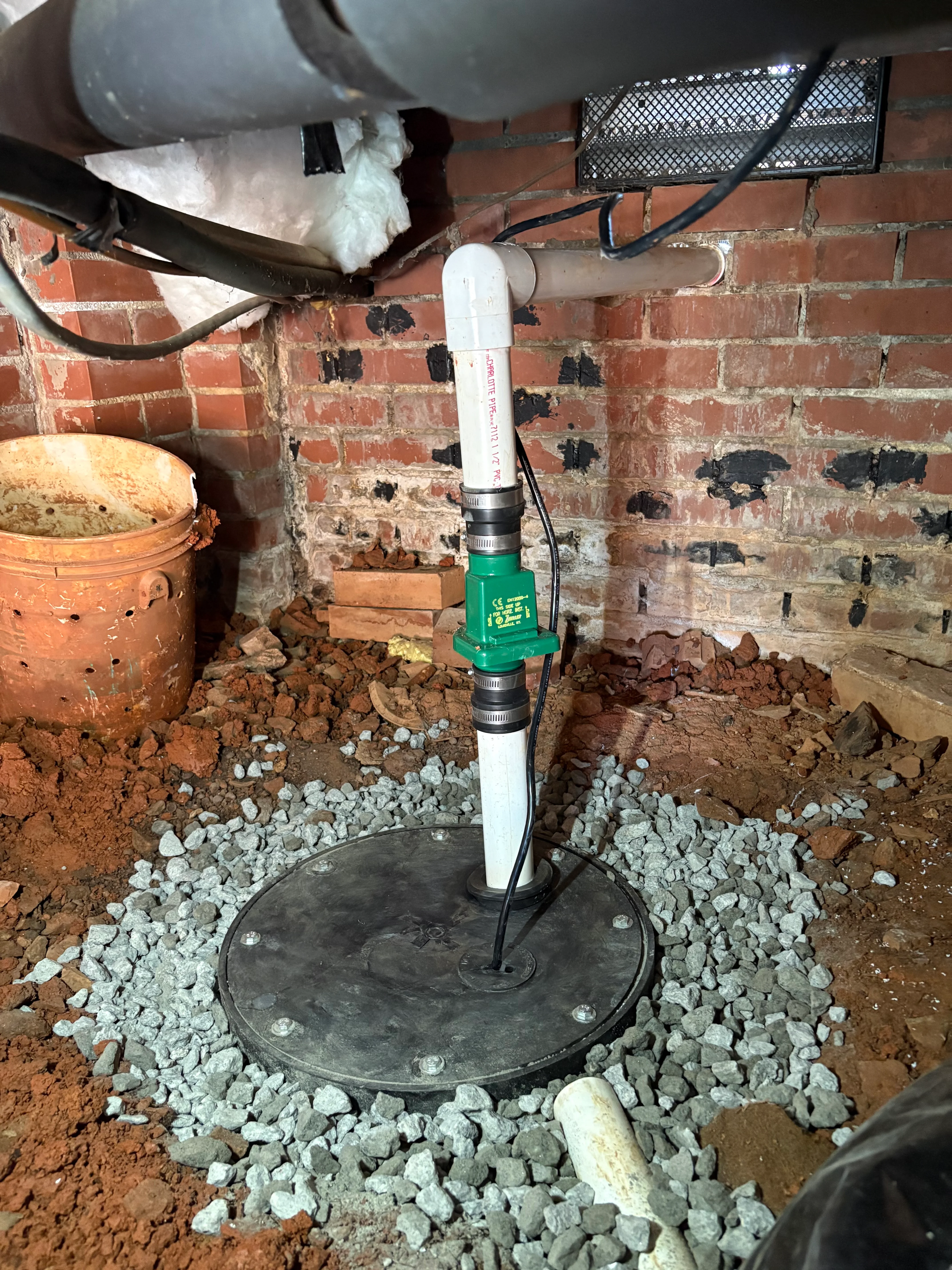 Installed sump pump with white discharge pipe and green valve in crawl space with gravel base