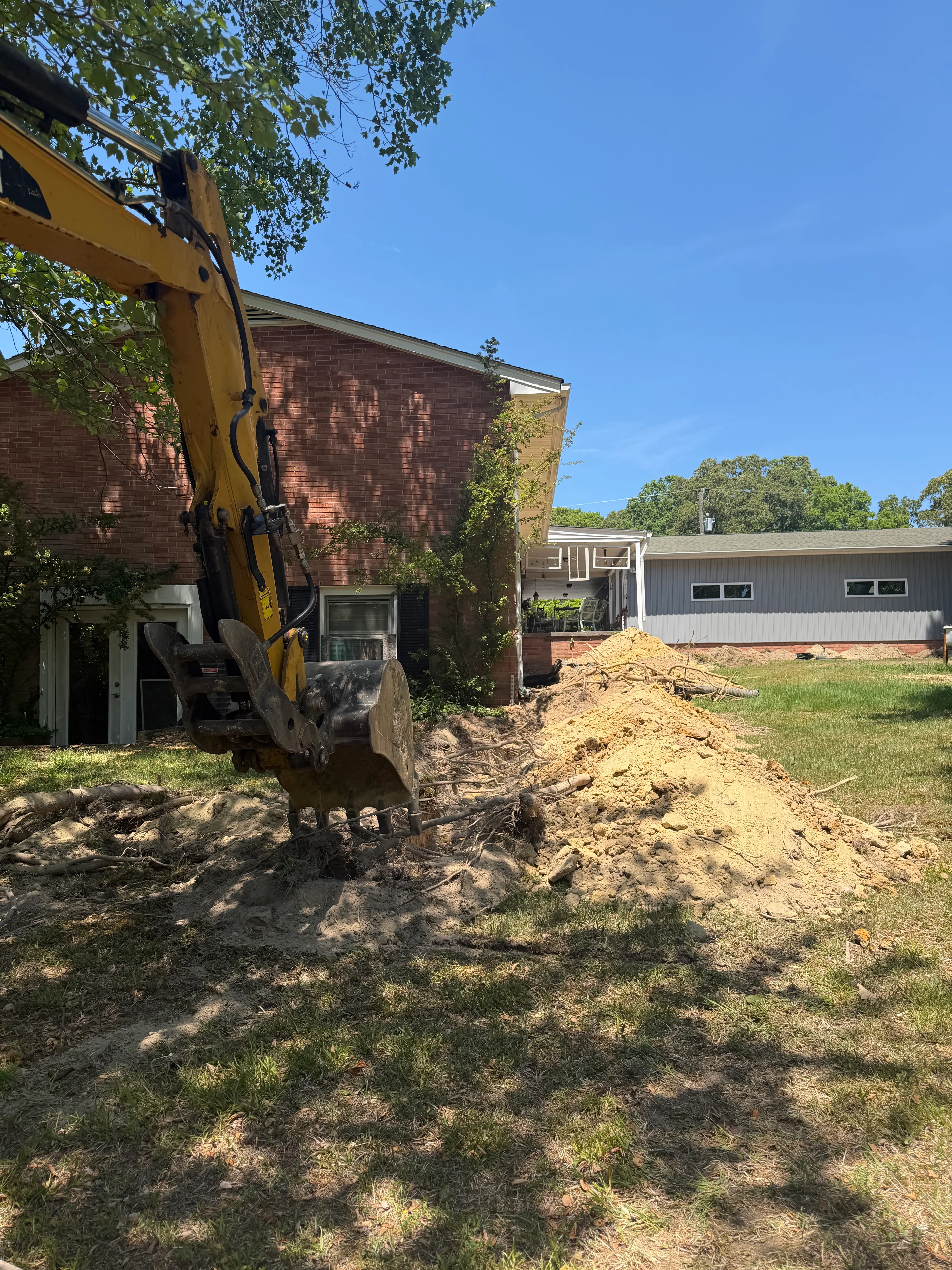 Yellow excavator removing soil from residential crawl space near brick home