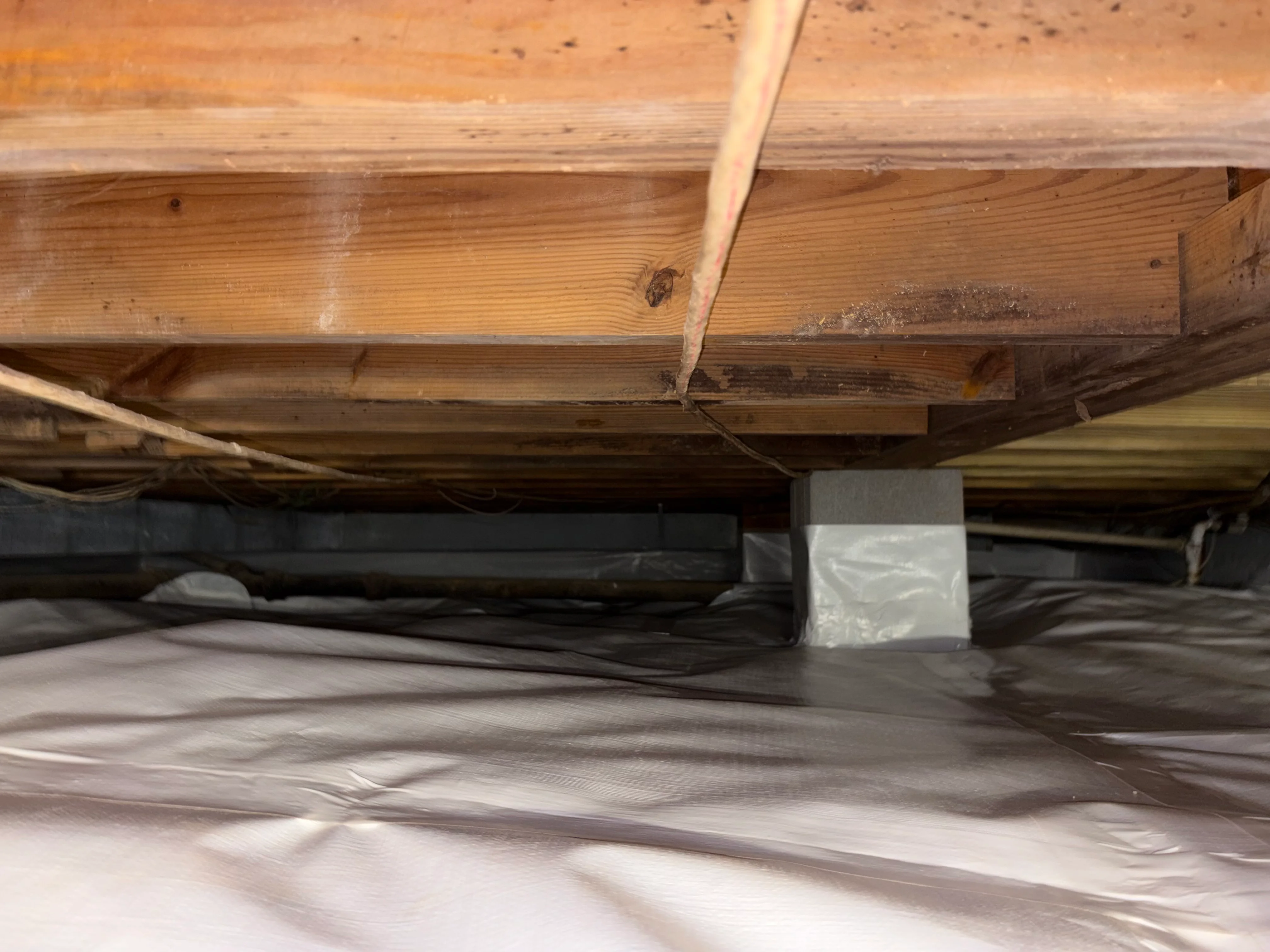 Crawl space interior showing wooden beams, concrete support post, and vapor barrier installation